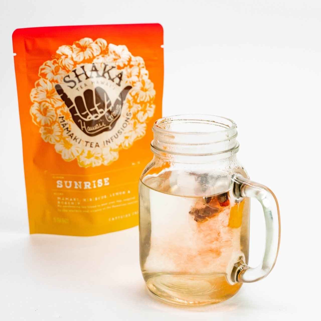 Shaka Sunrise tea package. Orange Tea brewing from tea bag in clear glass cup with glass handle. Mamaki, Hibiscus, lemon and rosehip